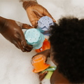 An adult and child play in soapy bathwater, holding Nuby Splish Splash Stacking Cups shaped like sea creatures. The child reaches for these colorful baby bath toys while bubbles cover their hands.