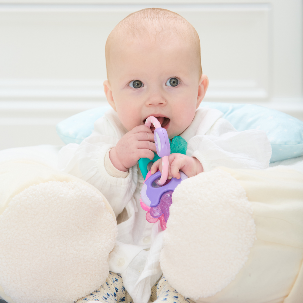 A baby with light skin sits in a plush seat, wearing a white outfit and holding the Icy Bite Keys Teether Toy while looking slightly upward.