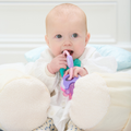 A baby with light skin sits in a plush seat, wearing a white outfit and holding the Icy Bite Keys Teether Toy while looking slightly upward.