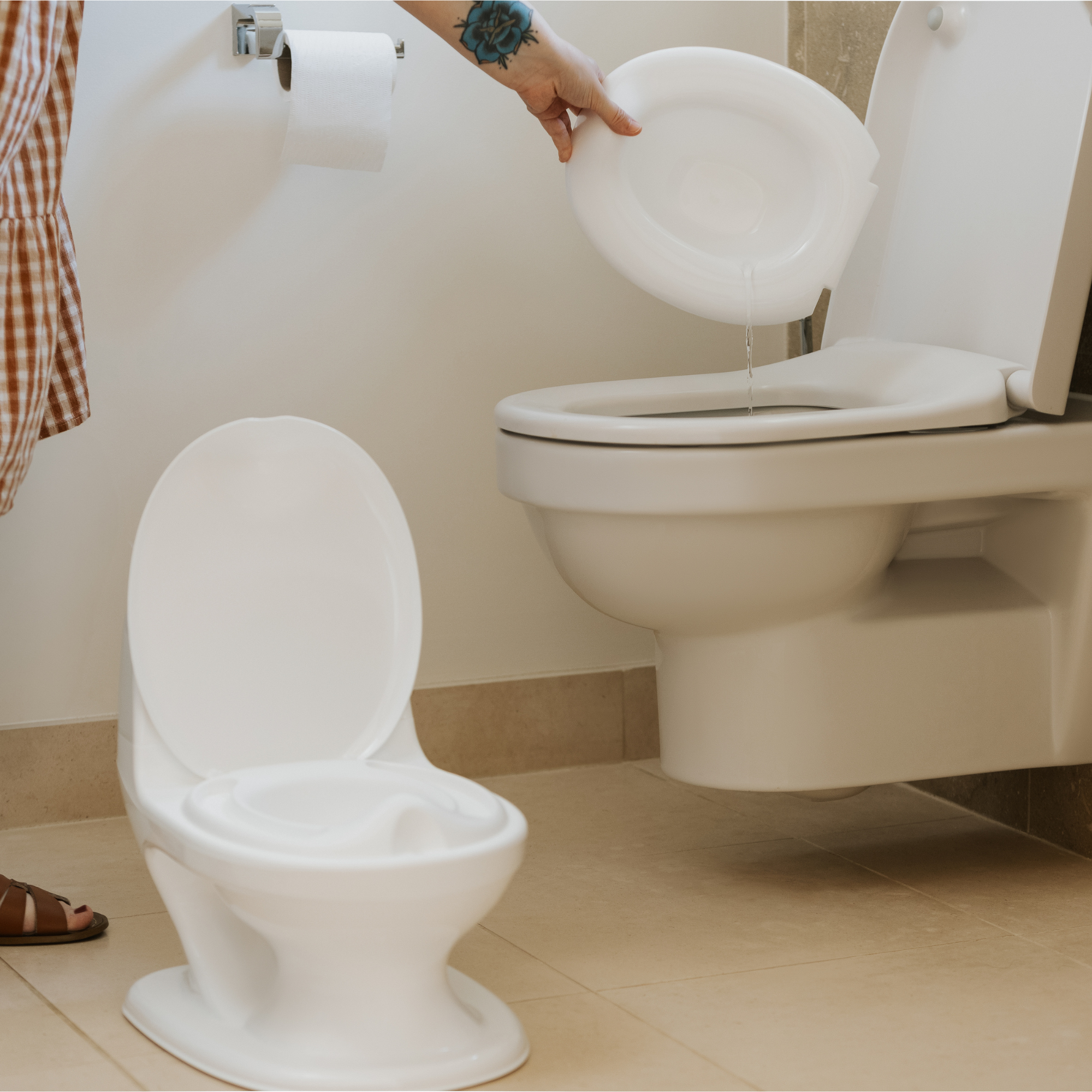 Someone empties a removable bowl from the white Nuby UK Potty Training Kit Bundle into a standard toilet. A roll of toilet paper is on the wall in this beige-tiled bathroom.