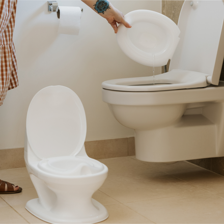 Someone empties a removable bowl from the white Nuby UK Potty Training Kit Bundle into a standard toilet. A roll of toilet paper is on the wall in this beige-tiled bathroom.