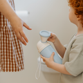 A child with curly hair holds a blue Nuby Gotta Go Travel Urinal as an adult reaches toward them in a bathroom, with a toilet and toilet paper holder visible in the background.