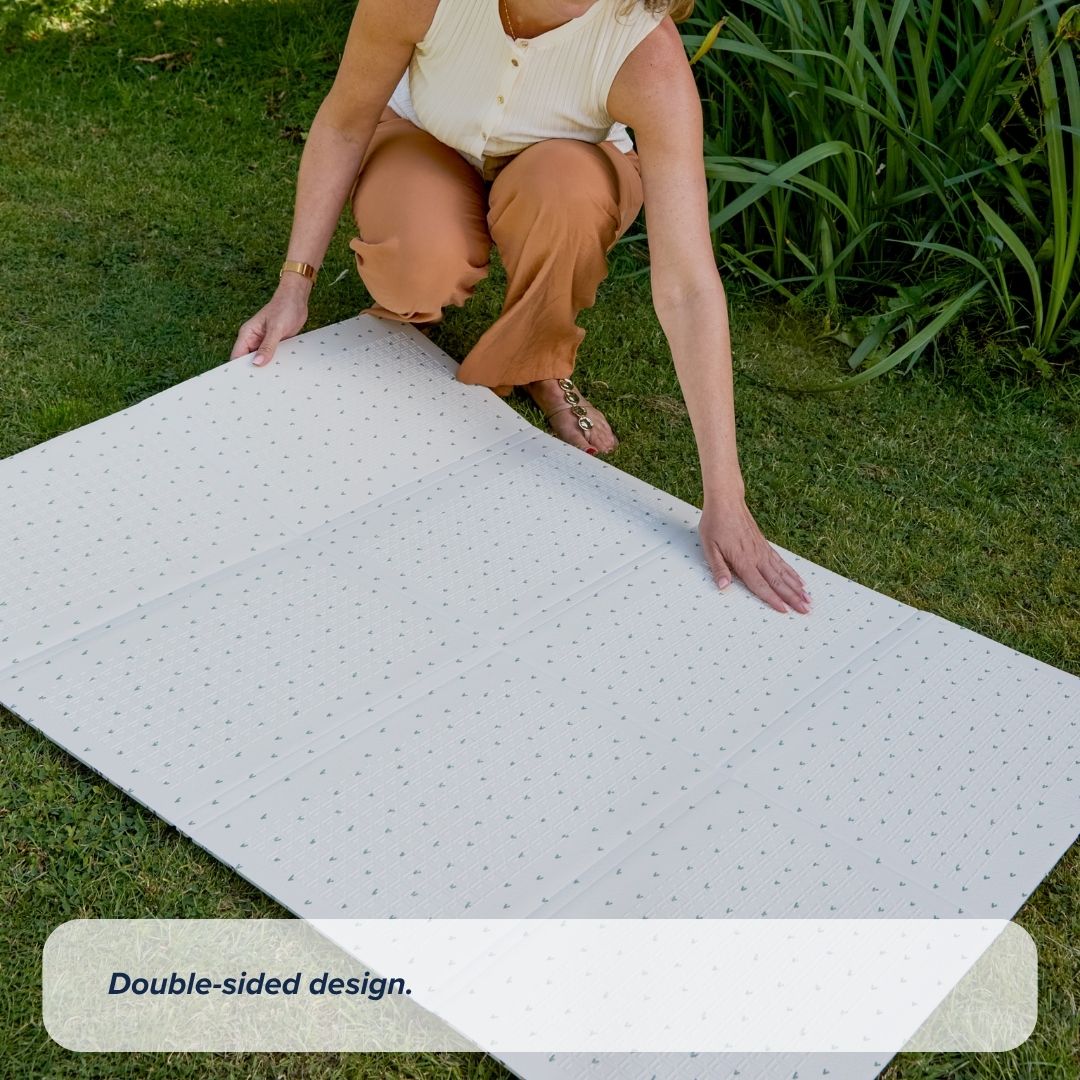 A woman kneels on grass, unfolding a large white Fold N Go Play Mat with a subtle pattern. Green plants grow nearby. Text at the bottom reads, Double-sided design.