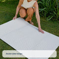 A woman kneels on grass, unfolding a large white Fold N Go Play Mat with a subtle pattern. Green plants grow nearby. Text at the bottom reads, Double-sided design.