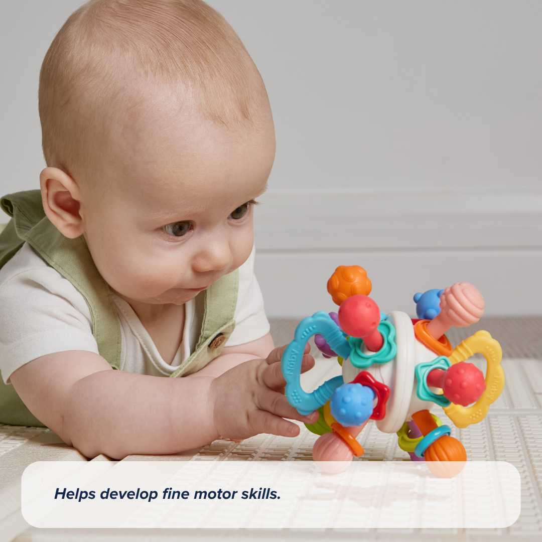 A baby in light overalls lies on a play mat, intently reaching for the Twist Ball Teether. Text on the image reads, Helps develop fine motor skills.