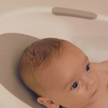 A close-up of a baby with wet hair lying in the Nuby Newborn Baby Bath, resting its head on a gray support and gazing up with wide eyes.