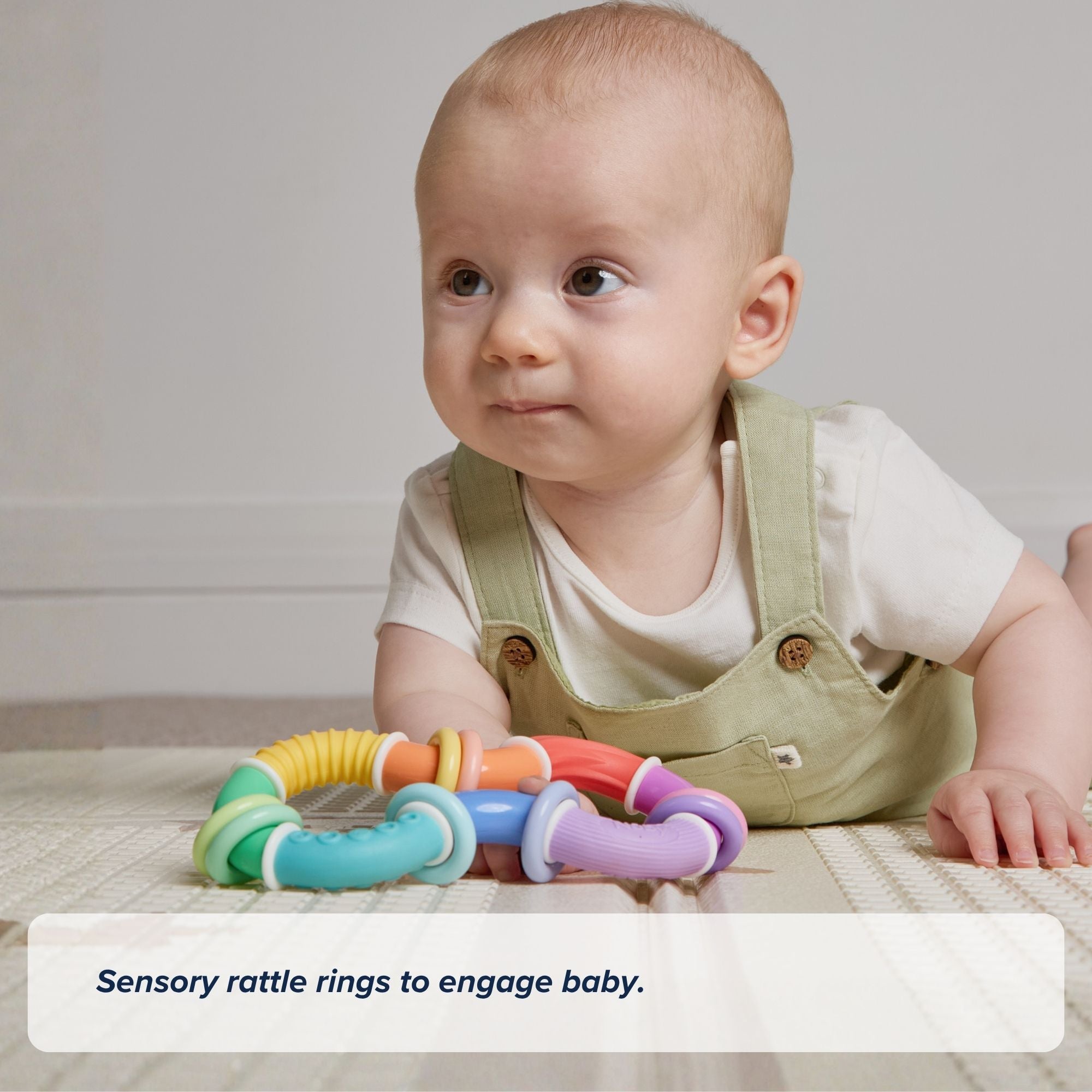 A baby in light green overalls lies on a mat, looking up, with the colorful Twista Rattle Teether placed in front of them.
