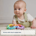 A baby in light green overalls lies on a mat, looking up, with the colorful Twista Rattle Teether placed in front of them.