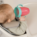 A baby in a high chair drinks from the Nuby All Around Cup 2 Pack - Pink, a spill-proof cup with a 360-degree valve, while wearing a light green sweater and secured with a padded safety harness.