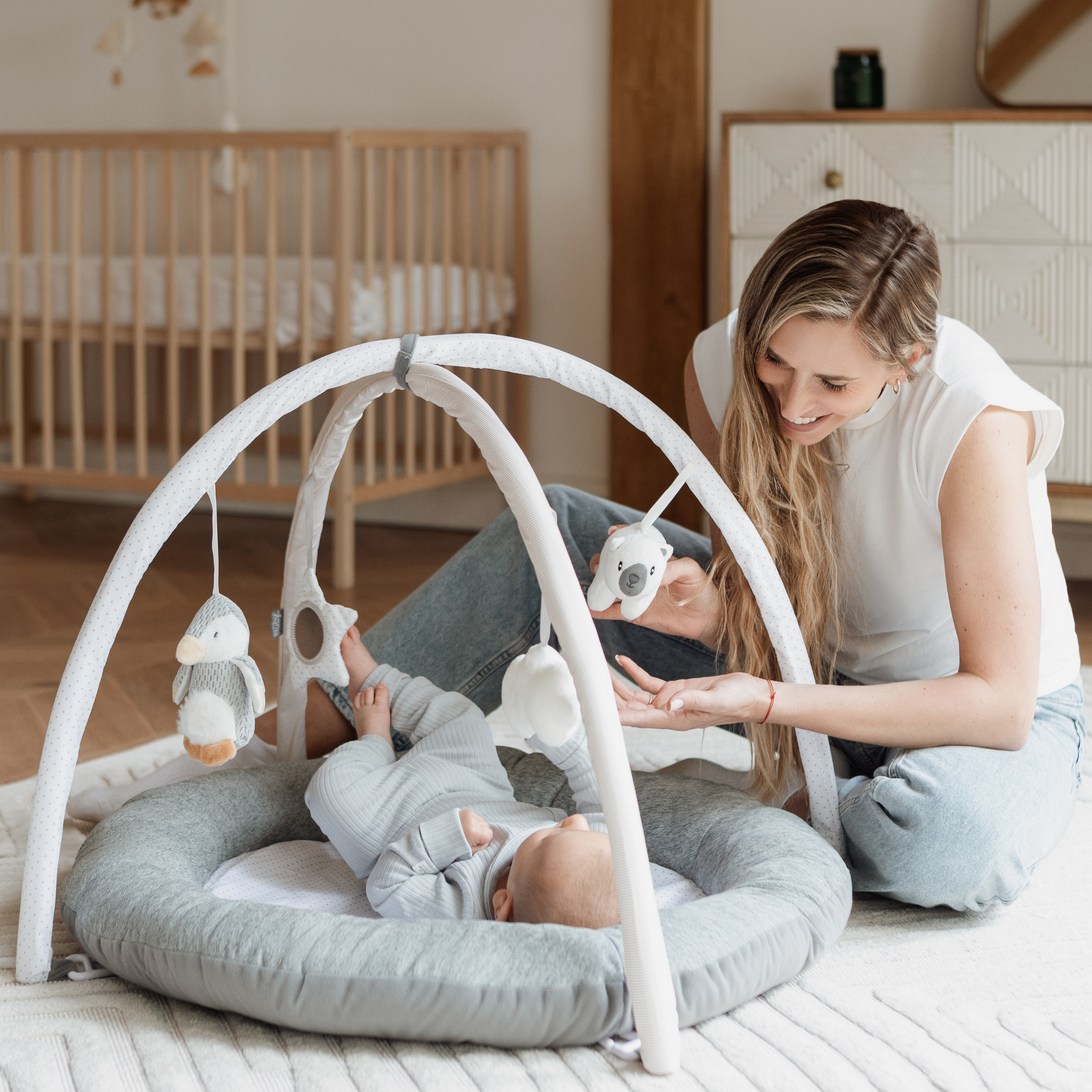 A woman sits on the floor next to a baby lying in a gray Nuby Penguin and Pals Play Gym, smiling and holding a hanging toy toward the baby in a cozy, modern nursery.