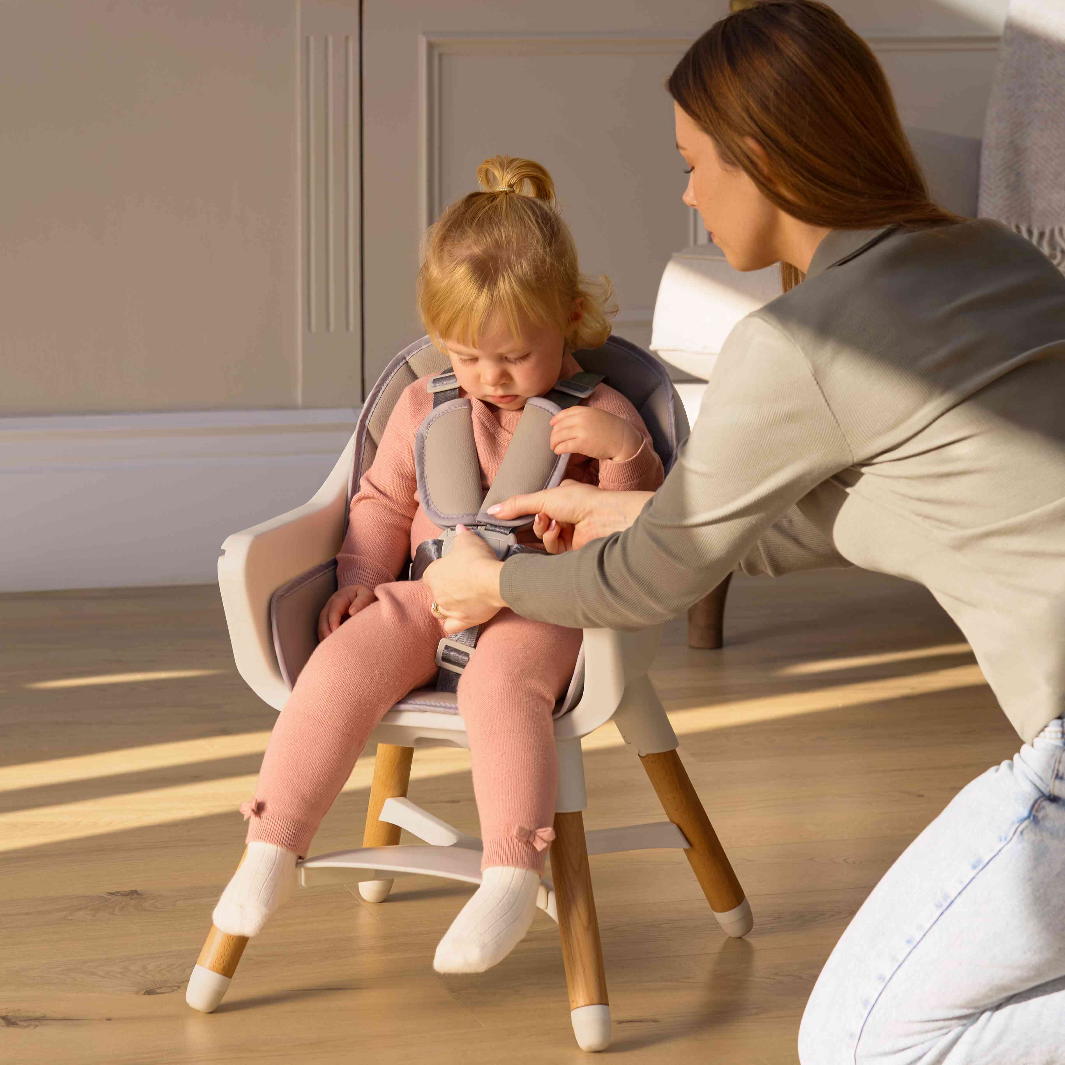 A woman fastens a safety harness on a young child in a Nuby UK 4-in-1 Multi-Use Highchair. The child, dressed in a pink outfit and white socks, sits as sunlight streams across the wooden floor.