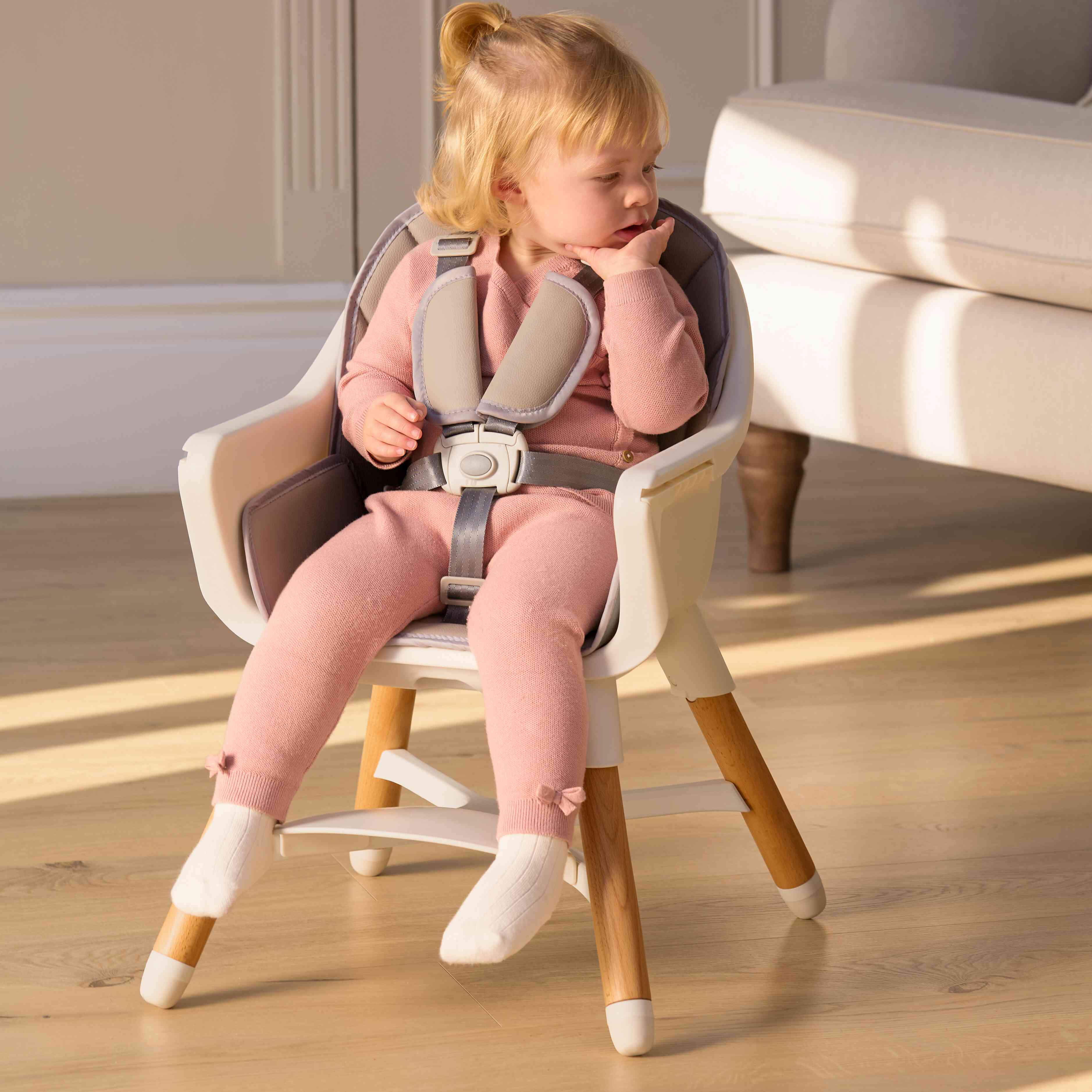 A young child with blonde hair sits safely strapped into the Nuby UK 4-in-1 Multi-Use Highchair, wearing a pink outfit and white socks, looking to the side as sunlight streams onto the wooden floor.