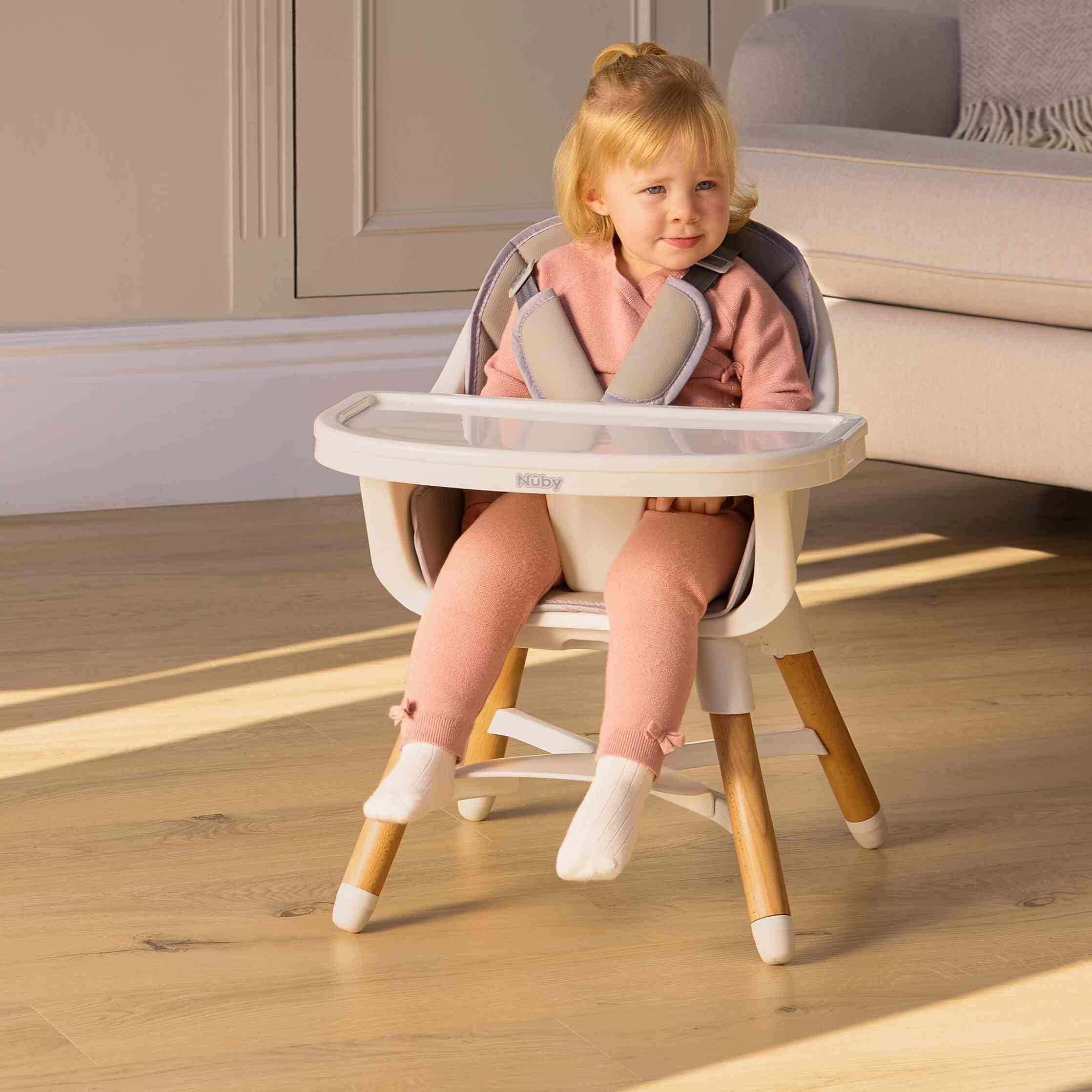 A young child sits in the Nuby UK 4-in-1 Multi-Use Highchair, dressed in pink and white socks. Sunlight brightens the wooden floor of a cozy, modern living room.