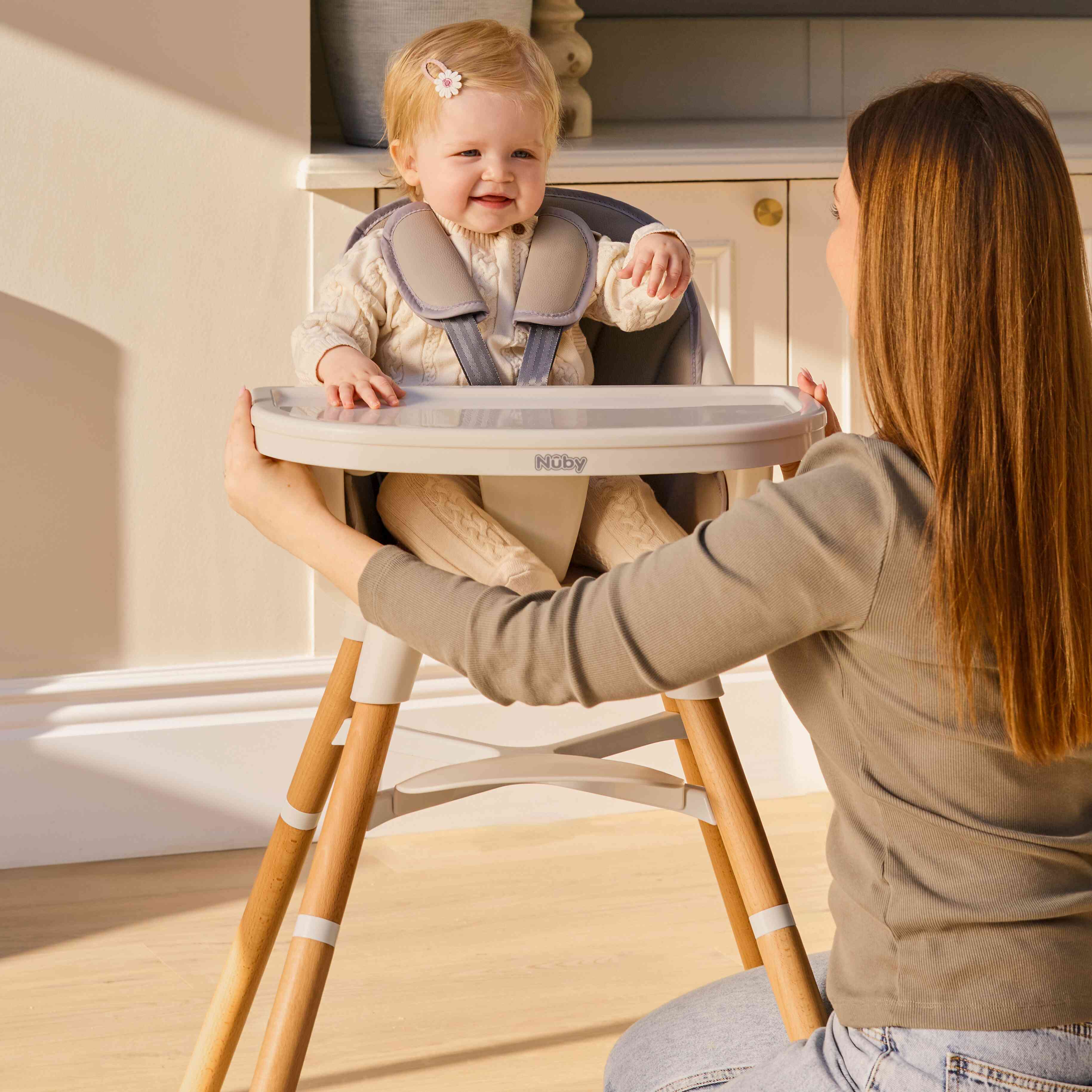 A woman kneels in front of a smiling baby dressed in cream with a hair clip, sitting in the Nuby UK 4-in-1 Multi-Use Highchair. She lovingly holds the sides of the convertible highchair as they share a joyful moment.