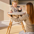 A woman kneels in front of a smiling baby dressed in cream with a hair clip, sitting in the Nuby UK 4-in-1 Multi-Use Highchair. She lovingly holds the sides of the convertible highchair as they share a joyful moment.
