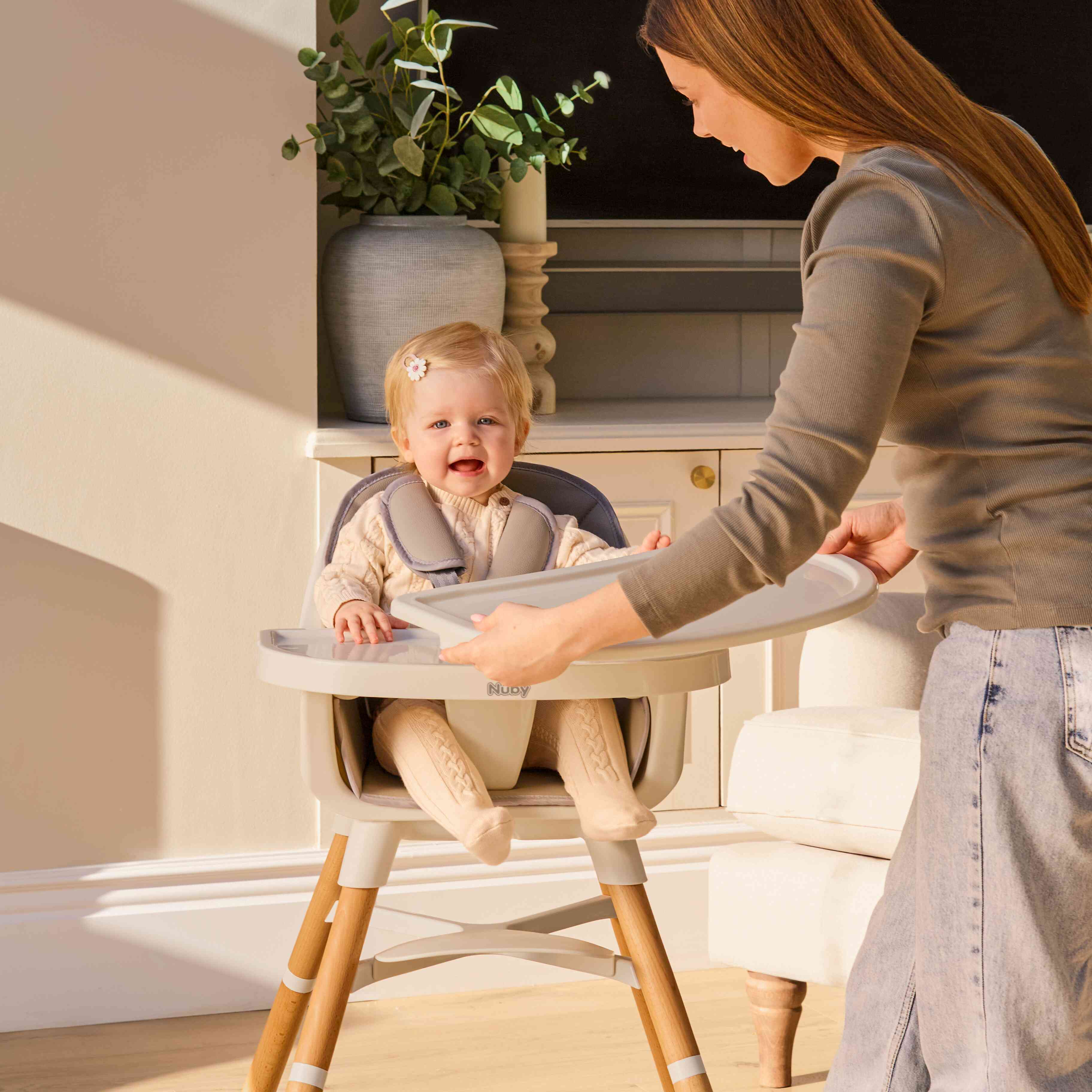 A woman attaches a tray to the Nuby UK 4-in-1 Multi-Use Highchair, where a smiling baby sits. Sunlight fills the room, with a plant and furniture visible in the background.
