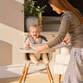 A woman attaches a tray to the Nuby UK 4-in-1 Multi-Use Highchair, where a smiling baby sits. Sunlight fills the room, with a plant and furniture visible in the background.