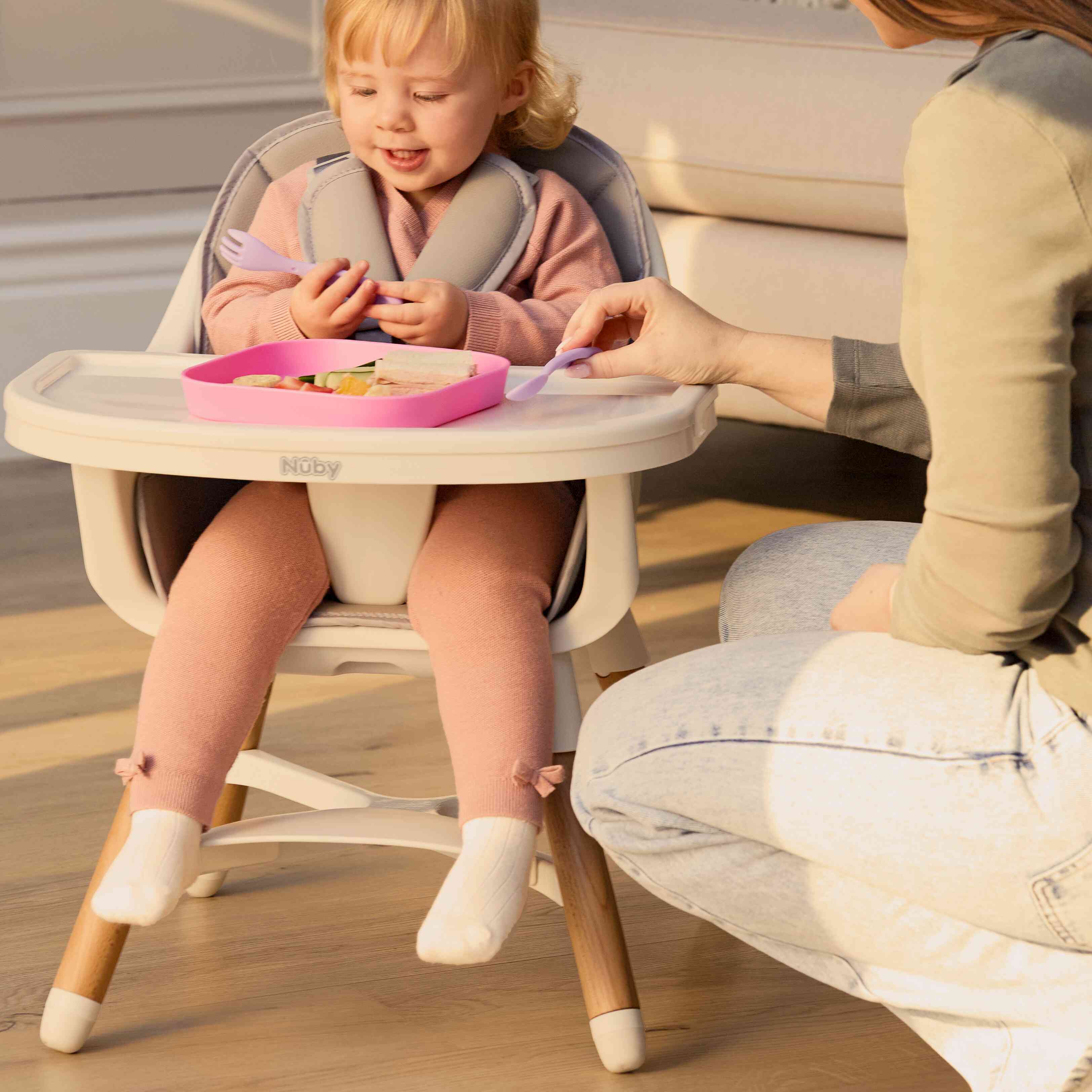 A toddler smiles in a Nuby UK 4-in-1 Multi-Use Highchair, holding a fork and spoon as an adult hands her a purple fork. A pink divided plate with food sits on the tray in front of her.