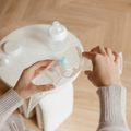 A person pours milk collected with the Nuby Silicone Manual Breast Pump into a storage bag, with baby bottles and lids on a round table nearby.
