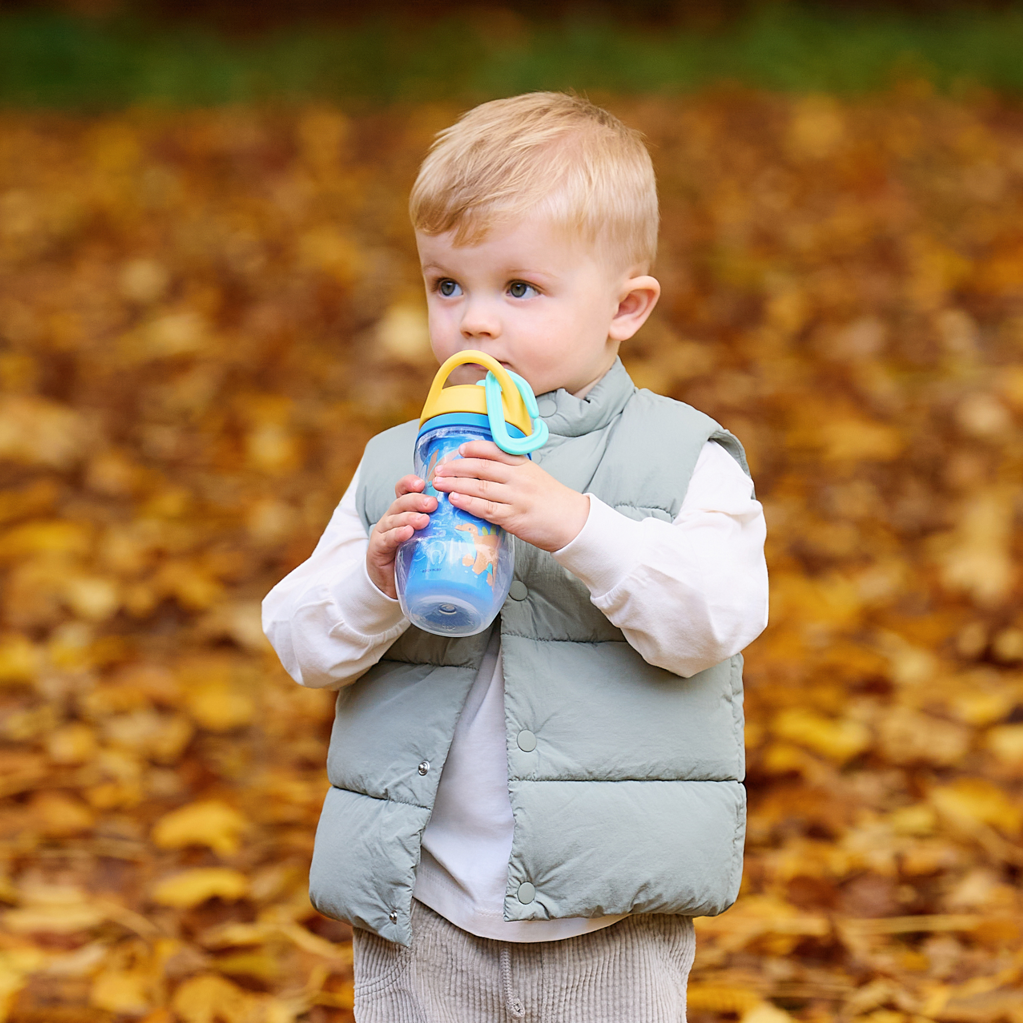 A young child with blonde hair, wearing a light blue puffer vest and holding an Insulated Sipper Cup 2 pack, stands outdoors on fallen autumn leaves.