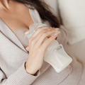 A person uses a Nuby Manual Breast Pump to express milk into a bottle while sitting, dressed in a light-colored robe or sweater.