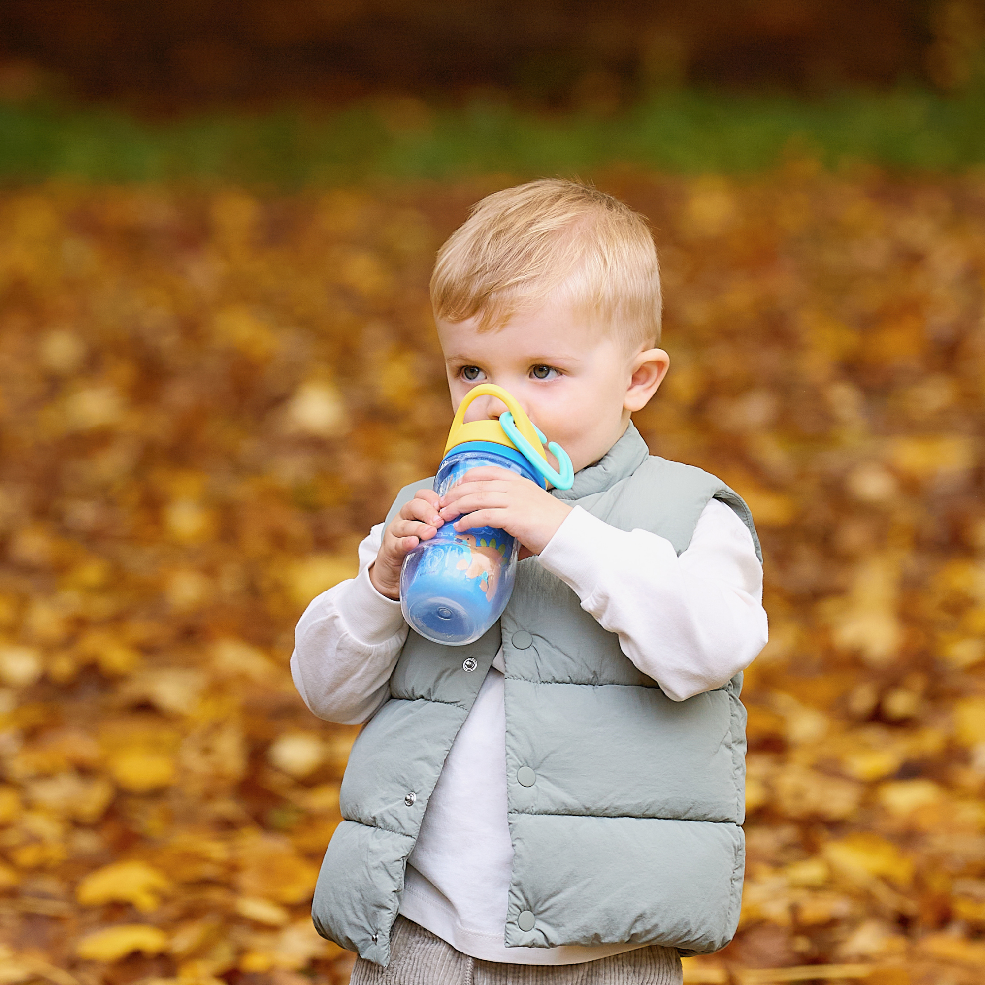 A young child wearing a light gray vest and white shirt stands outdoors on autumn leaves, drinking from the Insulated Sipper Cup 2 pack. The blurred background features soft yellow and brown tones.