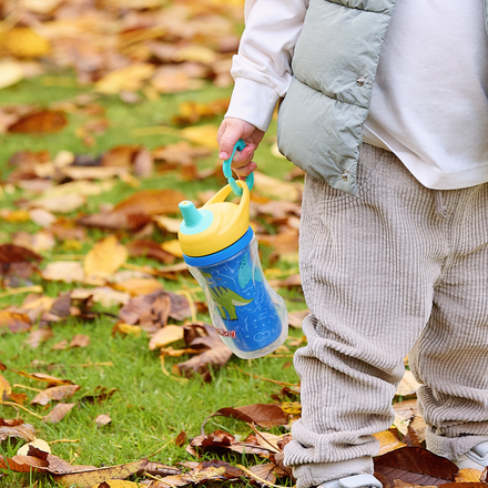 A young child in light pants, a white shirt, and a gray vest stands on autumn leaves holding an Insulated Sipper Cup 2 pack (blue and yellow), designed to be spill-proof for kids.