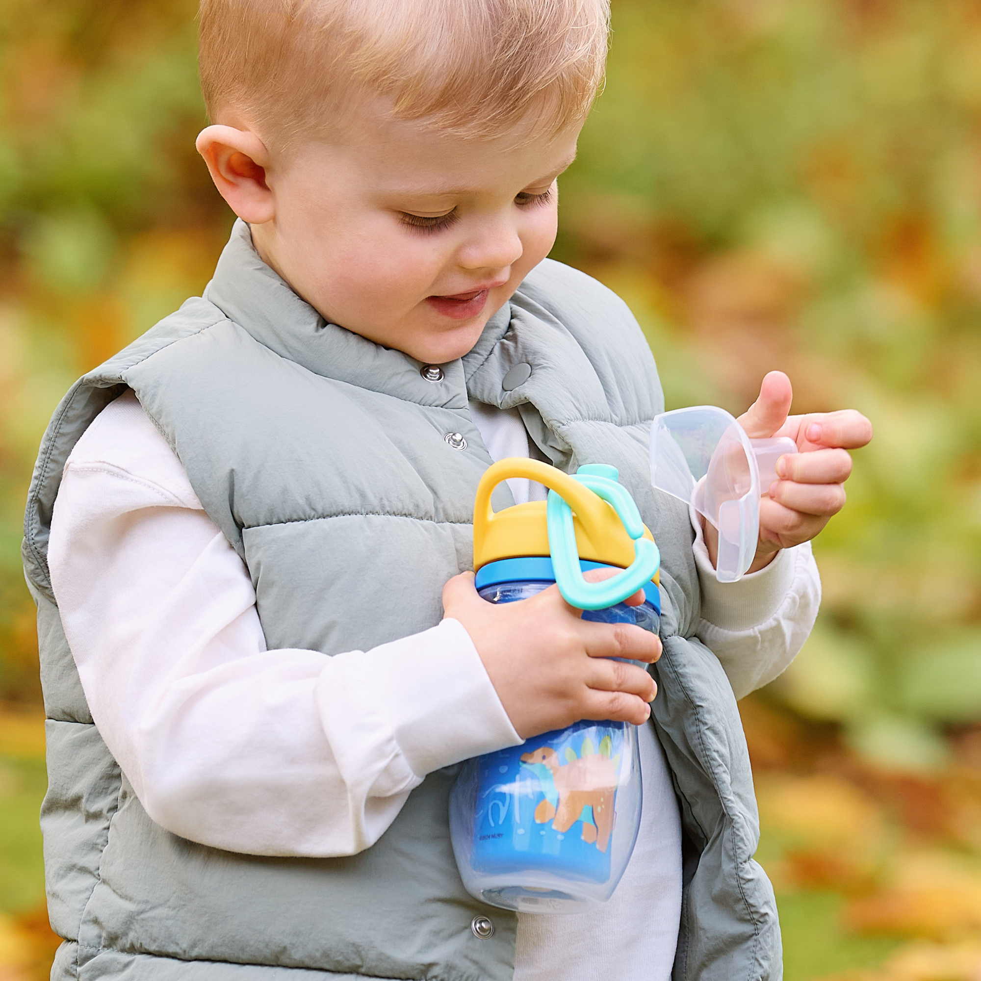 A young child in a light gray vest and white shirt holds an Insulated Sipper Cup 2 pack, removing the lid outdoors against a blurred backdrop of autumn leaves.