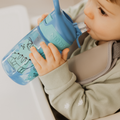 A young child drinks from a blue Nuby UK Incredible Gulp Water Bottle Dinosaurs, holding it with both hands while sitting in a high chair, wearing a light green shirt and gray bib.