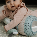 A baby in a pink ruffled sweater lies on a patterned play mat from the Nuby UK Animal Adventures Nursery Bundle, hugging a soft polka-dot animal toy and looking up with a gentle expression.
