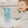 A smiling toddler sits in a high chair with a Nuby UK Incredible Gulp Water Bottle Dinosaurs, 4 Pack featuring a gentle silicone spout on the tray in front of them.