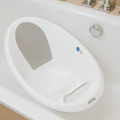 The Nuby Sit Me Up Baby Bath, featuring a gray backrest, raised support, and front handle for newborns, is placed in a white bathtub with a silver faucet and controls visible on the edge.