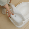 A child’s hand holds the handle of the Nuby My Real Mini Toilet Potty on a tiled floor, with part of the child’s legs and socks visible in the background.