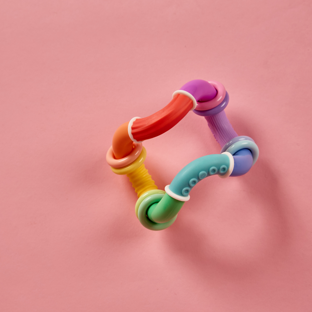 The Twista Rattle Teether is a colorful, flexible baby toy featuring multiple textures and segments arranged in a loose geometric shape on a pink background, providing soothing teething relief while also serving as a stimulating sensory toy.