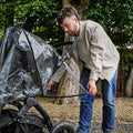 A man in a beige jacket adjusts a Pram Rain Cover on a black stroller outdoors, surrounded by trees and gravel.