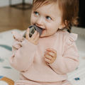 A toddler in a light pink sweater sits indoors, smiling and playfully exploring the Nuby Shape Sorter Learner Toy, which helps boost their hand-eye coordination.