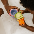 A child and adult play with Nuby Splish Splash Stacking Cups in a bubbly bath, pouring water between the colorful cups. Their hands and the cups are shown close up, surrounded by soapy bubbles.