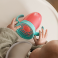 A baby in a high chair holds the Nuby All Around Cup 2 Pack - Pink with a 360-degree valve, examining the bottom. The baby is dressed in a soft, textured outfit and wears a gray bib.