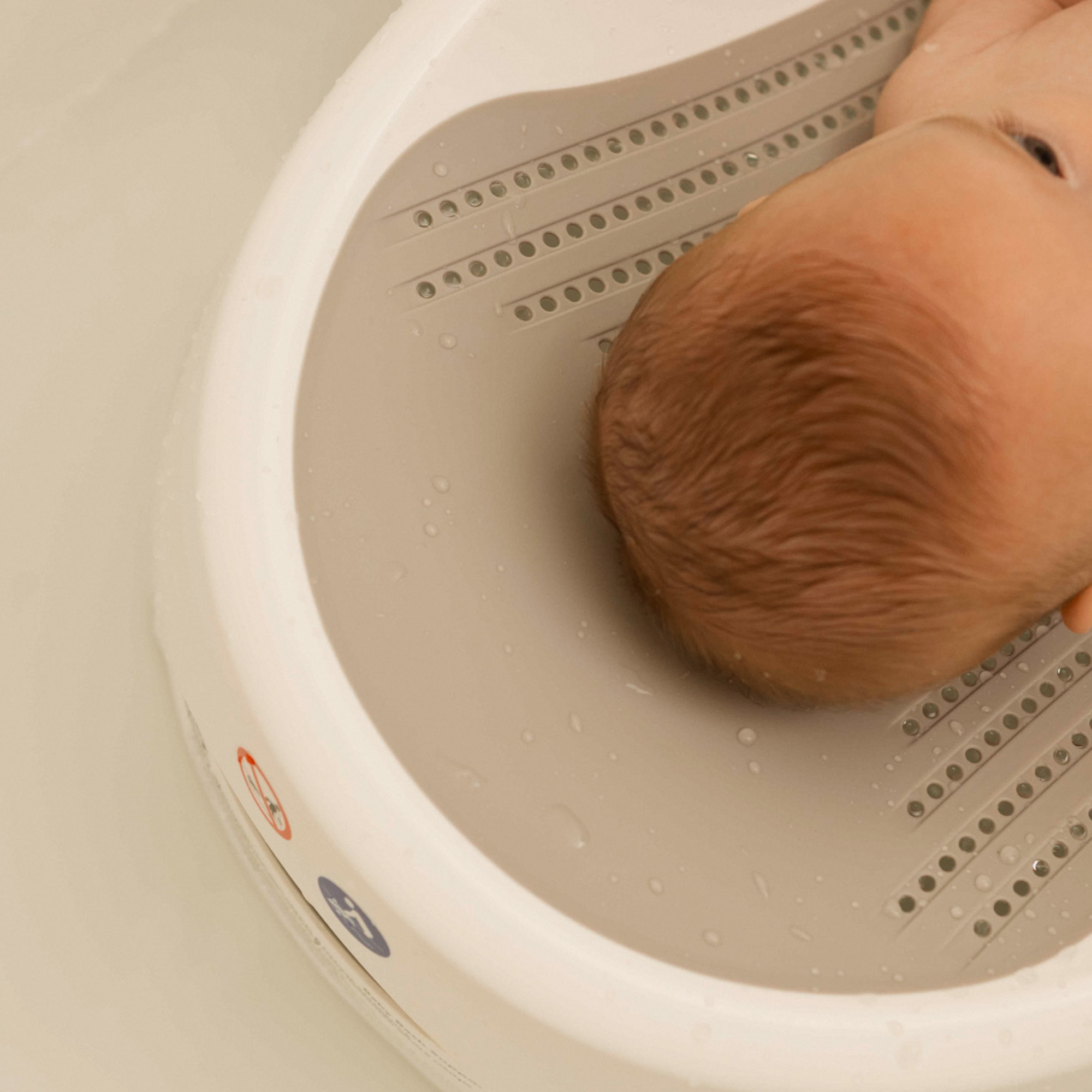 A baby with light brown hair rests in the Nuby UK Newborn Bath Support & Thermometer, a cushioned bath support fitting within a white plastic bath seat and partly submerged in water.