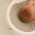 A baby with light brown hair rests in the Nuby UK Newborn Bath Support & Thermometer, a cushioned bath support fitting within a white plastic bath seat and partly submerged in water.