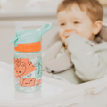 A Nuby UK Incredible Gulp Water Bottles 4 pack (Strawberries & Dinosaurs) sits on a highchair tray, while a smiling child in a light-colored bib is in the background.