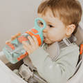 A toddler in a high chair drinks from a Nuby UK Incredible Gulp Water Bottle, part of a 4-pack featuring Strawberries & Dinosaurs designs. The child wears light green and is secured with a grey harness.