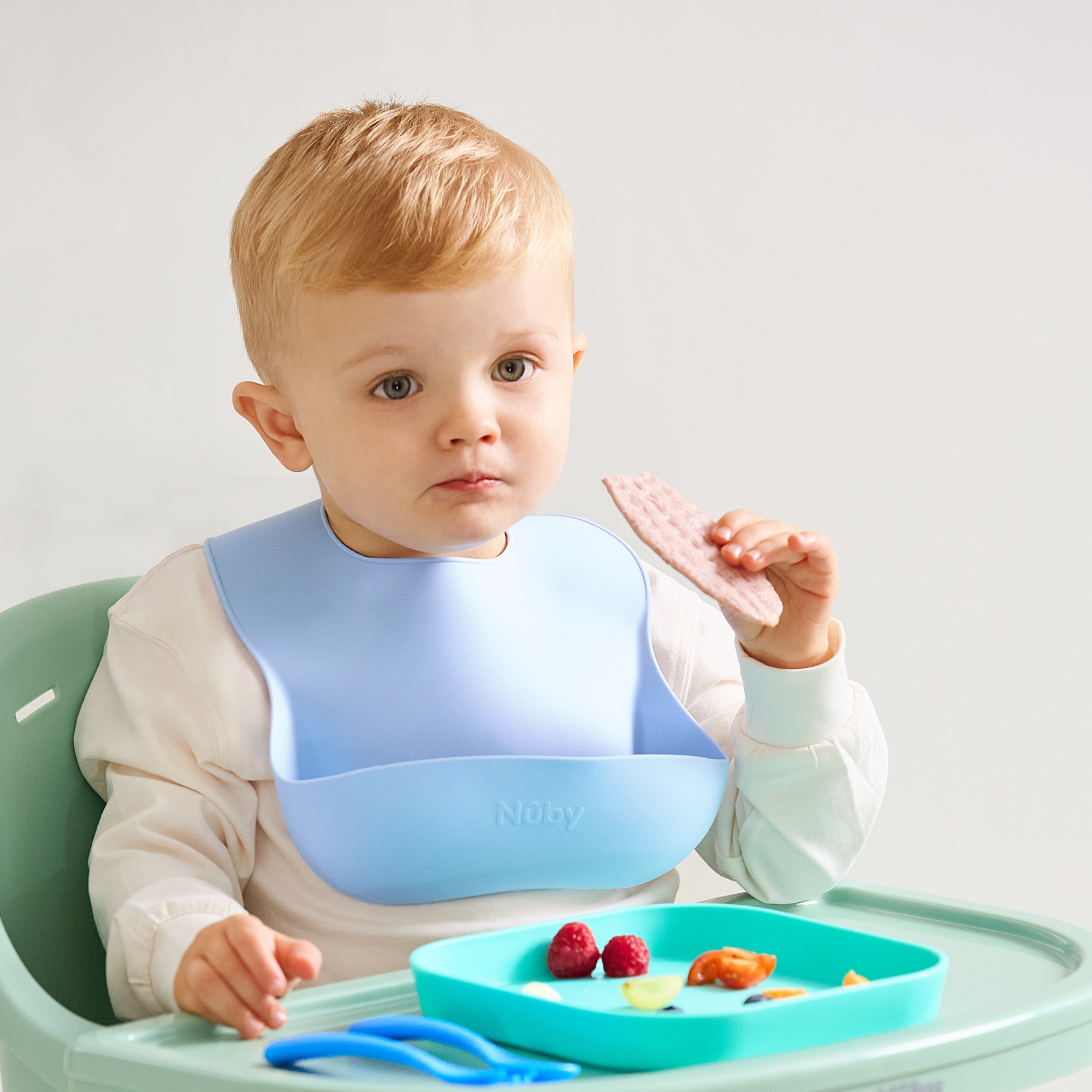 A toddler with short blond hair sits in a high chair, wearing a Silicone Bibs 3 Pack bib and holding a cracker. A blue plate with assorted food, like raspberries and vegetables, is on the tray in front of them.