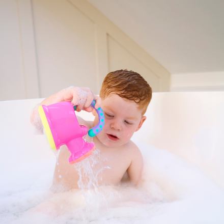 A young child with wet red hair sits in a bubble bath, pouring water from the Fun Watering Can, a bright pink and blue bath toy, surrounded by white foam.