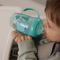 A young child in a high chair drinks from a turquoise Nuby UK Grip n Sip Cup (Whales & Bees, 4 Pack) decorated with sea animals. The child wears a light green outfit secured with a gray safety strap.