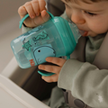 A young child in a sage green sweater sits in a high chair, drinking from a Nuby UK Grip n Sip Cup (Whales & Bees, 4 Pack). Turquoise cups are decorated with whales and other sea creatures.