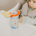 A baby sits in a high chair wearing a bib, with a Nuby UK Super Straw Twin Pack toddler cup featuring animal illustrations and a yellow lid with straw placed on the tray in front of them.