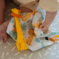 The Chompy Chube Teether in the design of yellow and orange forming the shape and look of a Lion. This is laid on top of an Animal Adventures product while being interacted with by a baby.