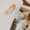 A child in a highchair holds a Nuby UK Super Straw Twin Pack cup—a clear sippy with animal designs, orange flip-top lid, and straw—offering a fun alternative to standard water bottles; Nuby is printed on the lid.