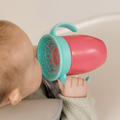 A baby in a high chair, wearing a light green sweater, drinks with both hands from a pink Nuby All Around Cup 2 Pack.